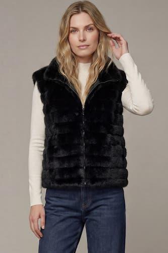 A woman modeling a black faux fur zip-up vest over a cream long-sleeve top and dark blue jeans, set against a solid light gray studio background.