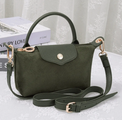 An olive green faux suede handbag with a top handle, flap closure, and adjustable shoulder strap, styled on a white lace tablecloth with a book in the background.
