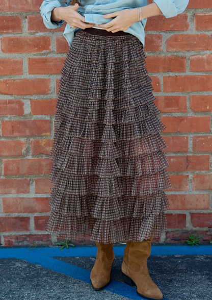 A person wearing a Brown plaid mesh maxi skirt with tiered ruffle layers and flowing silhouette, styled with a light blue shirt and tan boots, standing in front of a red brick wall.