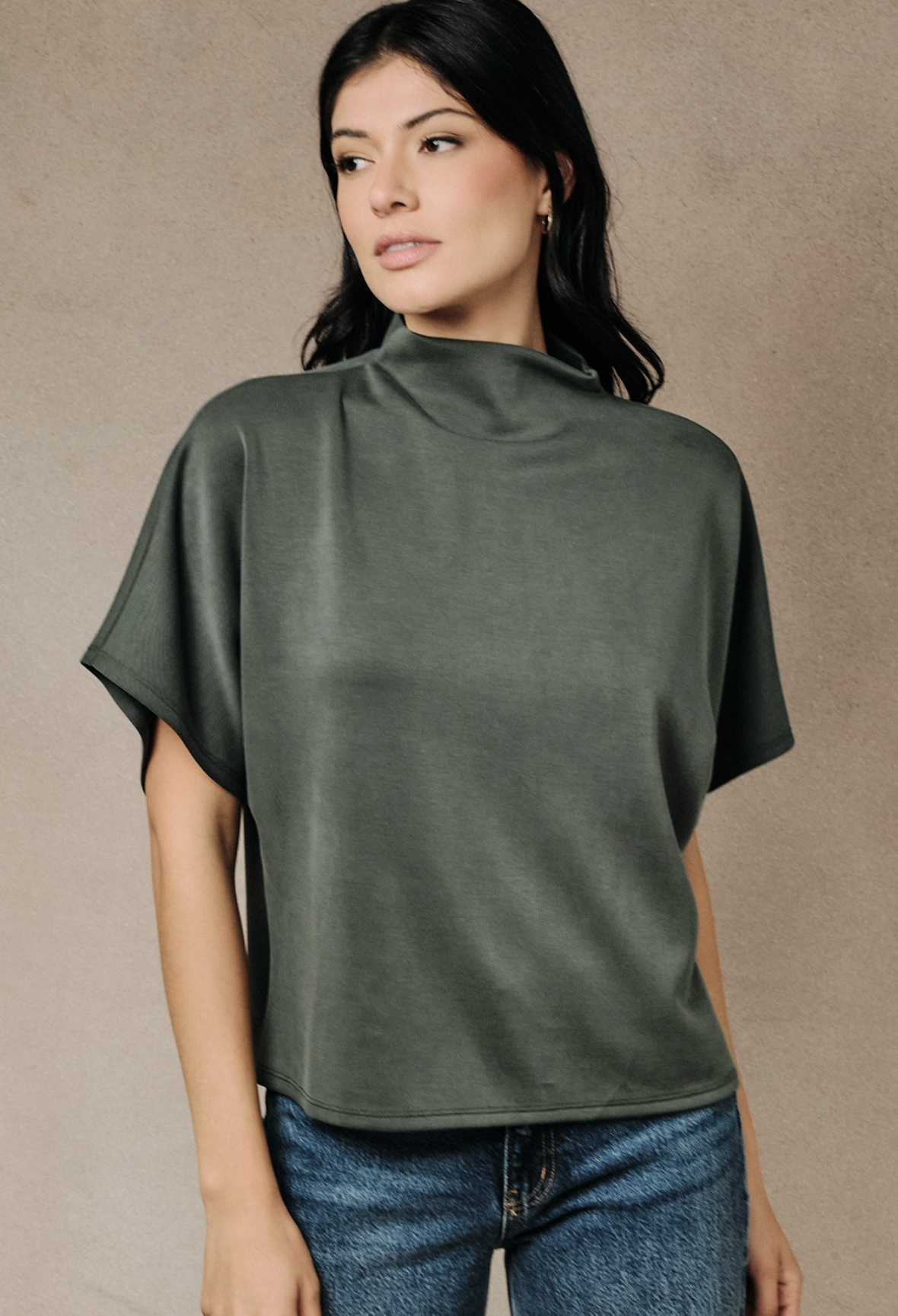 A woman wearing and Olive dolman short sleeve top with lightweight drape and relaxed silhouette and blue jeans, standing against a textured beige studio backdrop.
