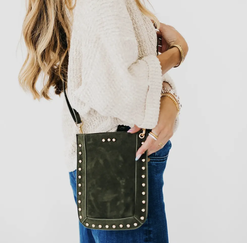 A woman carrying a dark green crossbody bag with silver studs and a dark green strap against a plain white background.