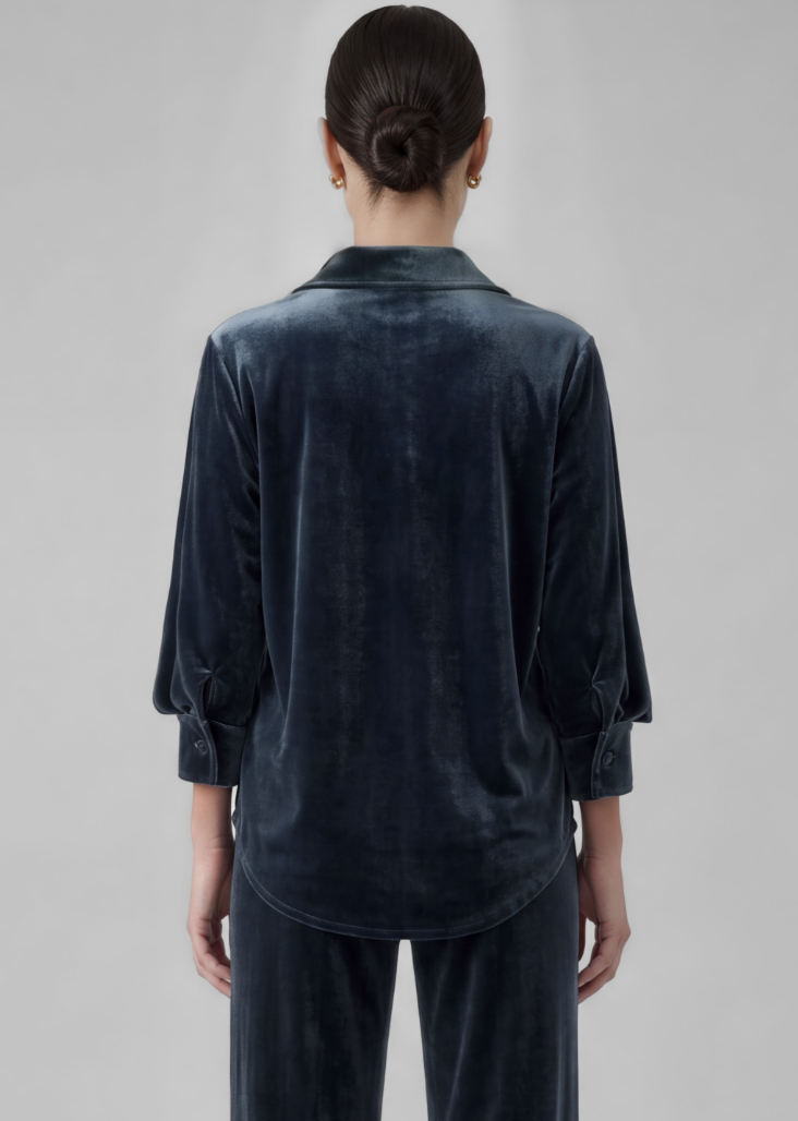 A woman viewed from the back wearing a dark navy velvet long-sleeve collared shirt and matching dark pants against a light gray studio background.