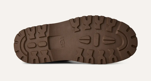 A close-up view of a brown rubber shoe sole with deep tread patterns, showing the embossed 'UGG' logo, on a light off-white background.