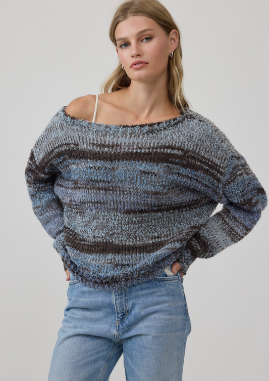 A woman wearing a chunky knit off-the-shoulder sweater with horizontal stripes in heathered blue, gray, and dark brown, paired with light-wash blue jeans, against a plain light gray studio background.