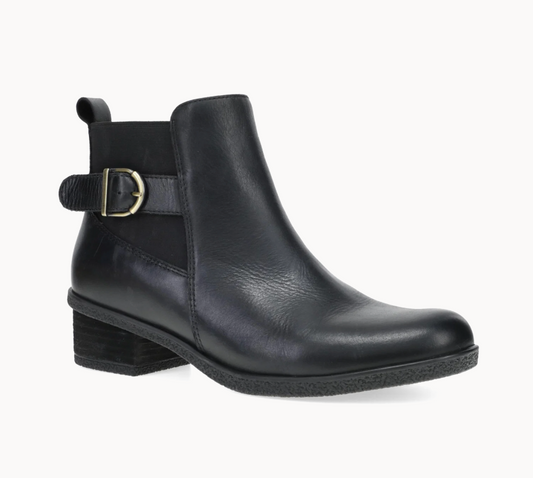 A black leather Dansko Deedee ankle boot with a side strap and gold buckle, shown from a three-quarters angle on a light gray background.