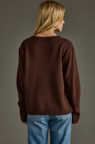 A woman shown from the back wearing a dark brown knit sweater and blue jeans against a solid dark gray studio background.