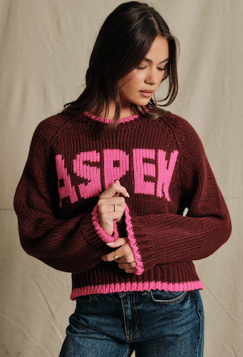 A woman wearing a dark burgundy ribbed knit sweater with bright pink lettering that reads 'ASPEN' across the chest, paired with blue jeans, standing against a neutral beige fabric backdrop.