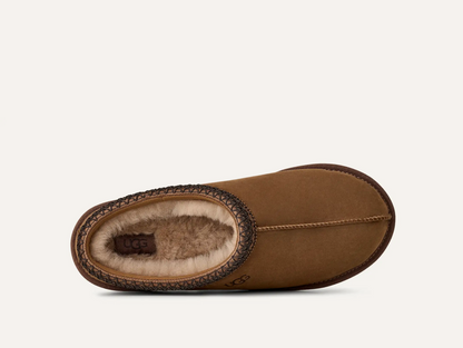 A single UGG slipper shown from a side profile view against a light off-white background. The slipper features a warm brown suede exterior, a dark brown braided trim, and a plush cream-colored shearling lining.