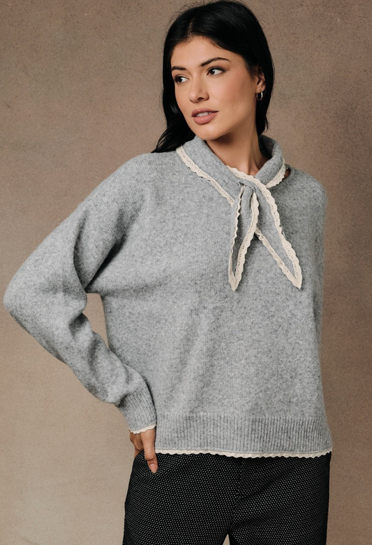 A woman wearing a Heather gray scarf‑neck sweater with relaxed silhouette and soft brushed knit, paired with black patterned pants, set against a textured neutral studio background.