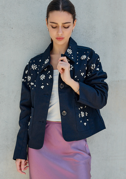 A woman wearing a cropped navy blue jacket with white floral embroidery on the shoulders and upper chest, styled over a white top and a lavender satin skirt, standing against a textured gray concrete wall.