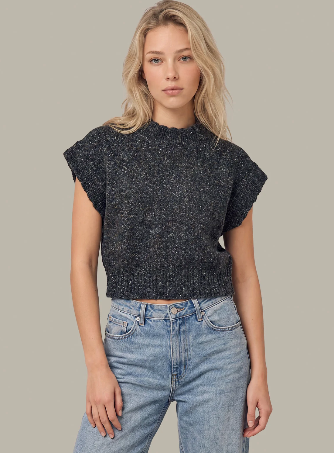A blonde woman wearing a dark charcoal gray textured 525 America knit short-sleeve crop sweater and light wash blue jeans, standing against a solid neutral gray background.