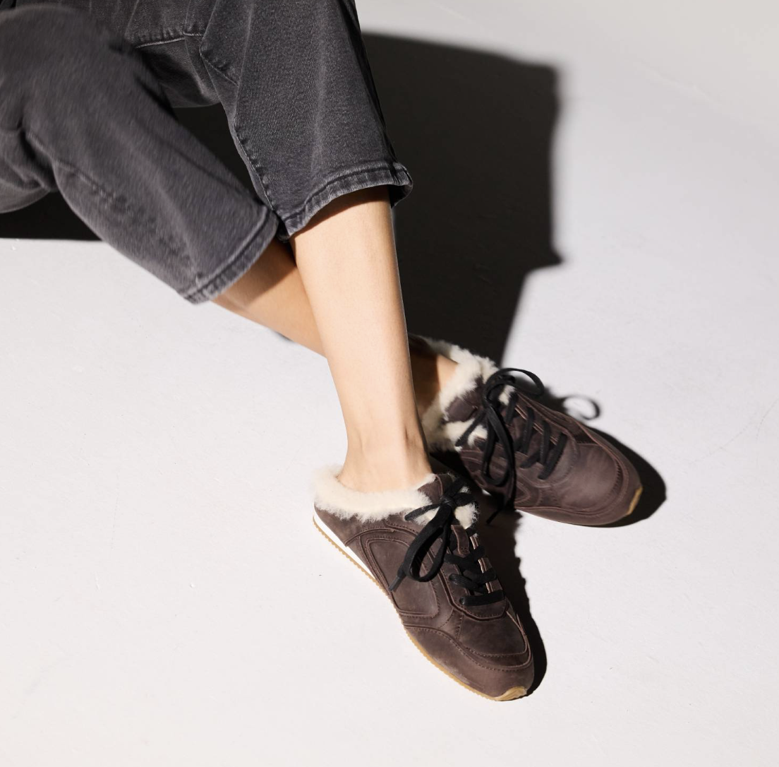 A person seated on a white floor wearing Yosi Samra dark brown suede sneakers with white faux-fur trim and black laces, paired with dark gray jeans. 