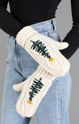 A person wearing cream knit winter mittens with green tree designs and small yellow accents, paired with light blue jeans and a black long-sleeve top against a gray studio background.