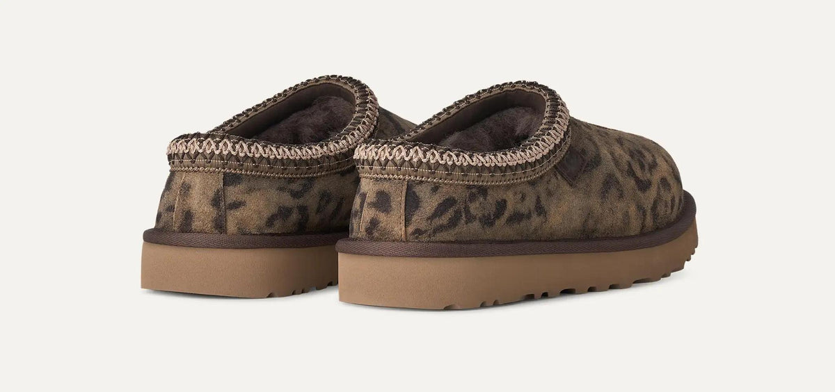 A pair of UGG Tasman Spotted clog slippers with a brown and black leopard print pattern, featuring a brown faux-fur lining and a thick brown sole, shown against a light off-white background.