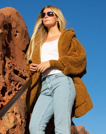 A woman wearing a brown sherpa Nordic Beach fleece wrap, white tank top, and light blue jeans stands outdoors against a clear blue sky and a large reddish rock formation.