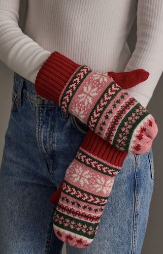 A person wearing a white ribbed sweater and blue jeans holds a pair of knit winter mittens with a red, pink, and dark green Fair Isle pattern.