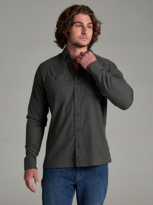 A man wearing a Kuhl Revolvr  dark charcoal gray long-sleeve button-down shirt and blue jeans, posing against a solid gray studio background.