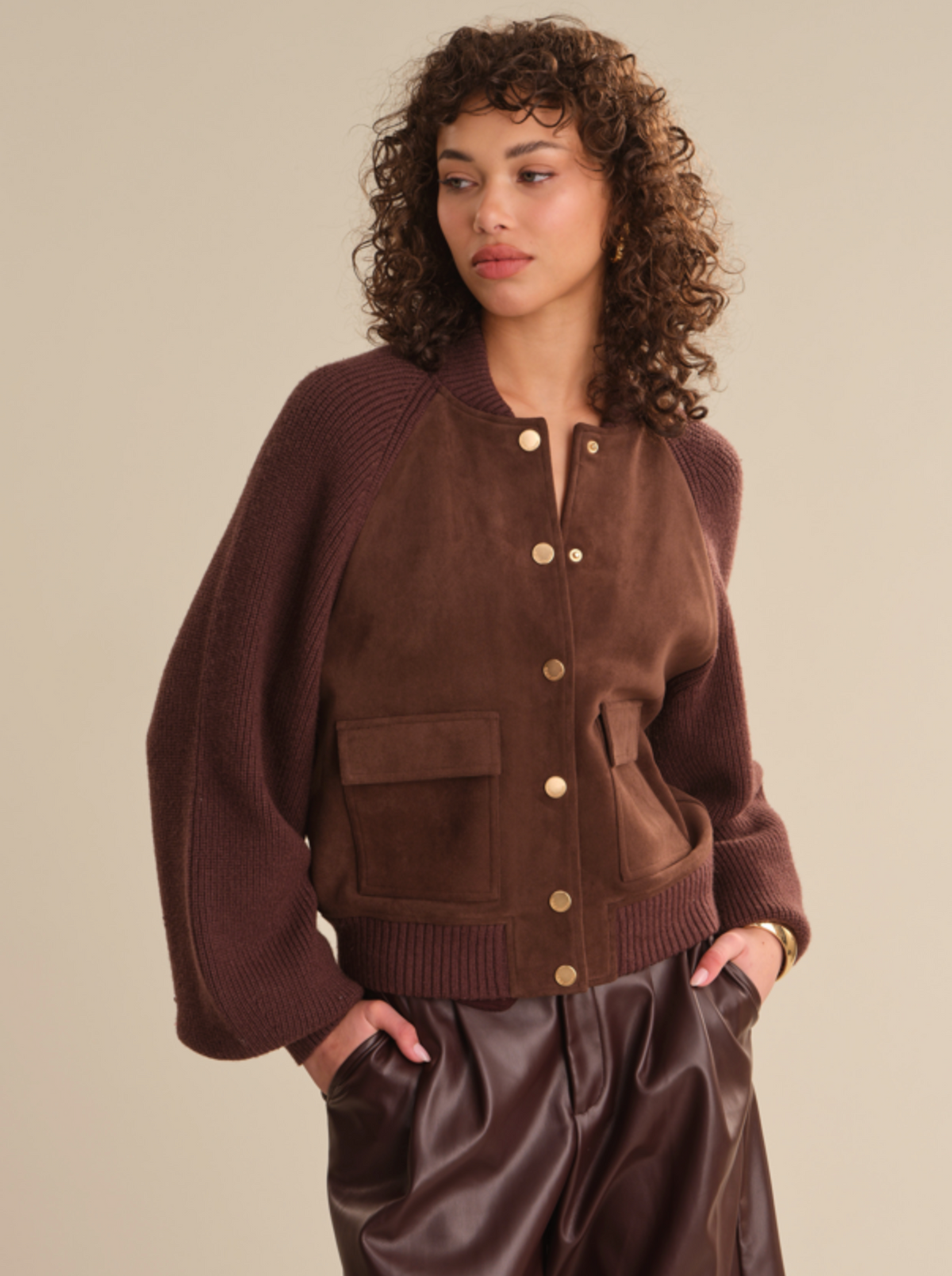 A woman wearing a brown suede and knit raglan bomber jacket with gold buttons, paired with dark brown faux-leather pants, against a solid beige background.