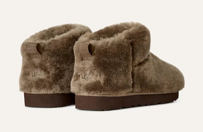 A pair of UGG Classic Ultra Mini ankle slipper boots in taupe brown faux fur, shown from a three-quarters rear angle against a light off-white background.