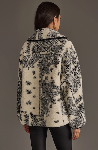 A woman viewed from the back wearing a cream sherpa-style jacket with a black paisley pattern and black trim, paired with black leggings, against a dark gray studio background.