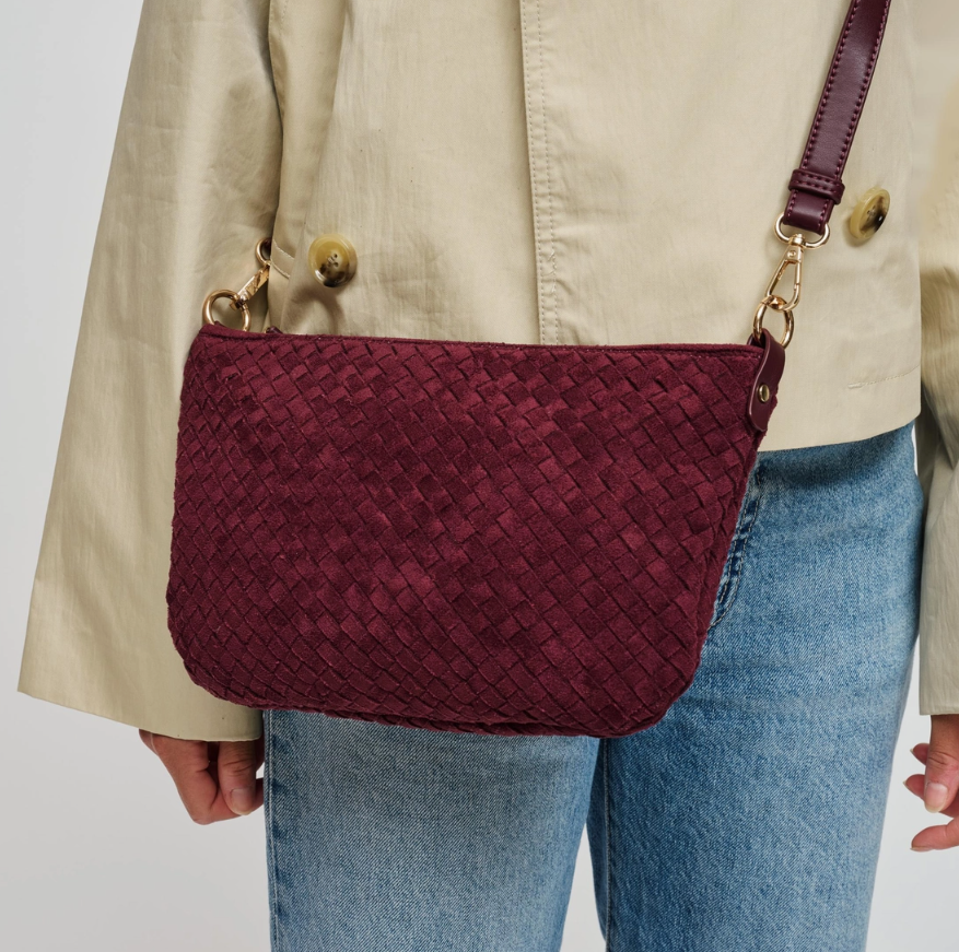 A burgundy woven leather crossbody bag with gold-tone hardware, worn by a model wearing a beige jacket and blue jeans.