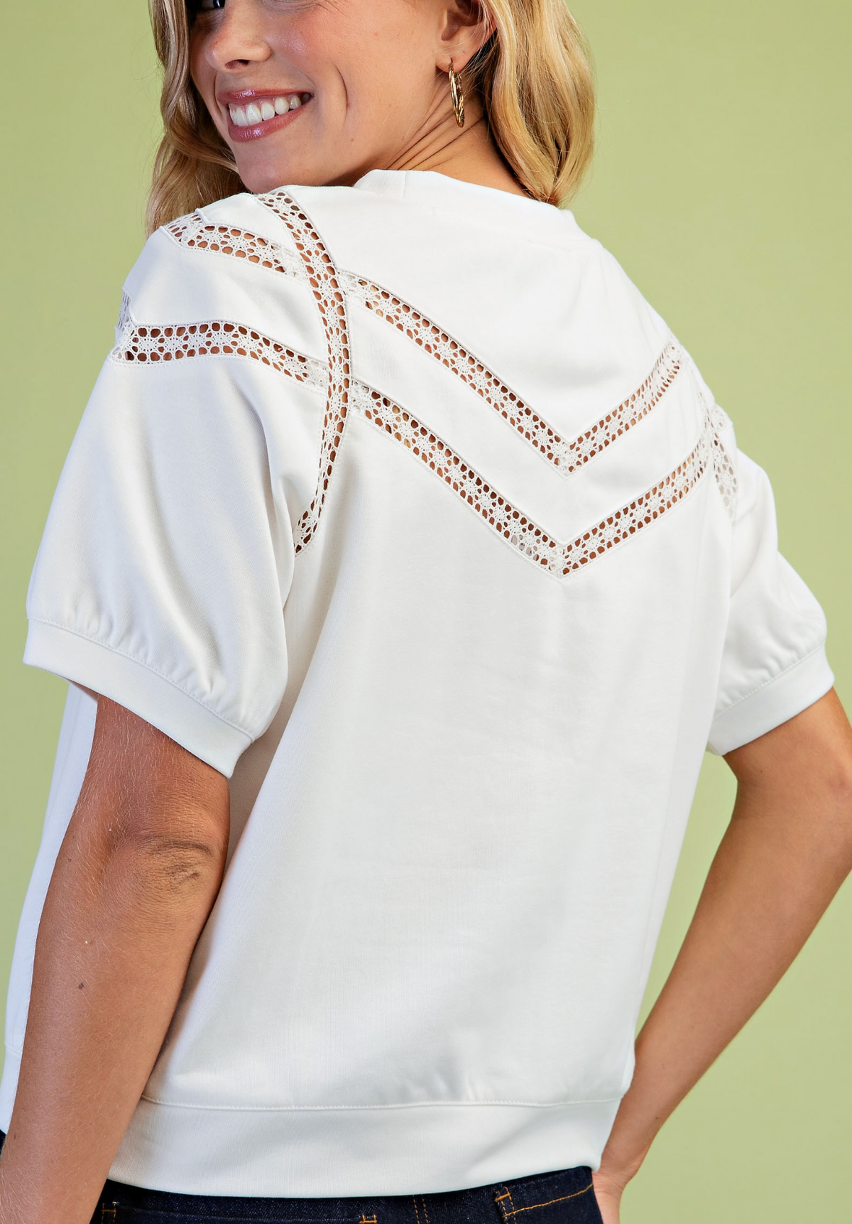 A woman wearing a white short-sleeve top with decorative brown crochet lace embroidery across the upper back, shown from a rear three-quarter angle against a solid light green background.