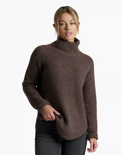 A woman wearing a brown Kuhl Sienna ribbed knit turtleneck sweater and dark jeans, standing against a plain white studio background.