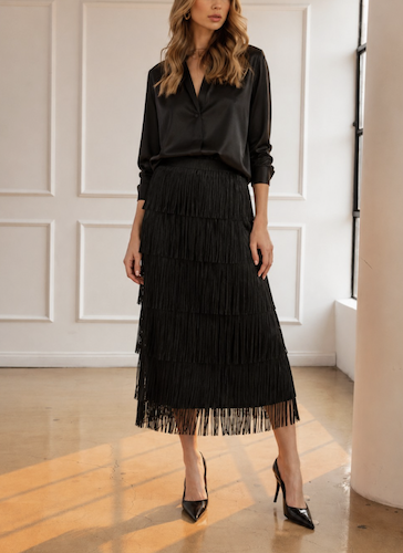 A woman models a black tiered fringe midi skirt paired with a black satin blouse and black pointed-toe heels, standing in a bright room with white paneled walls and a polished wood floor.