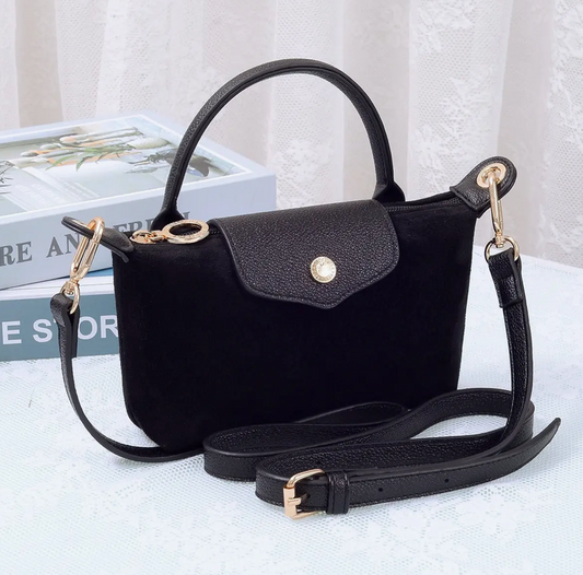 A black suede handbag with a top handle, flap closure, and detachable shoulder strap, styled on a white textured surface with books in the background.