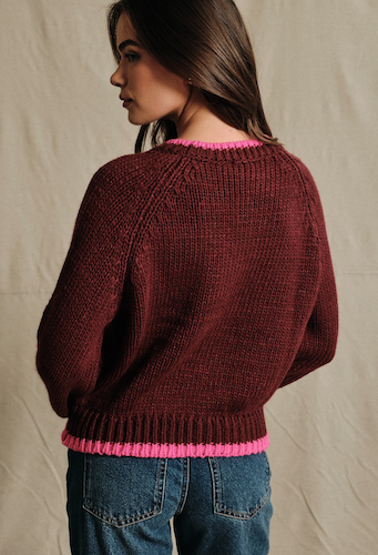 A woman shown from the back wearing a chunky knit sweater in deep burgundy with bright pink ribbed trim at the collar and hem, paired with blue jeans against a neutral fabric backdrop.