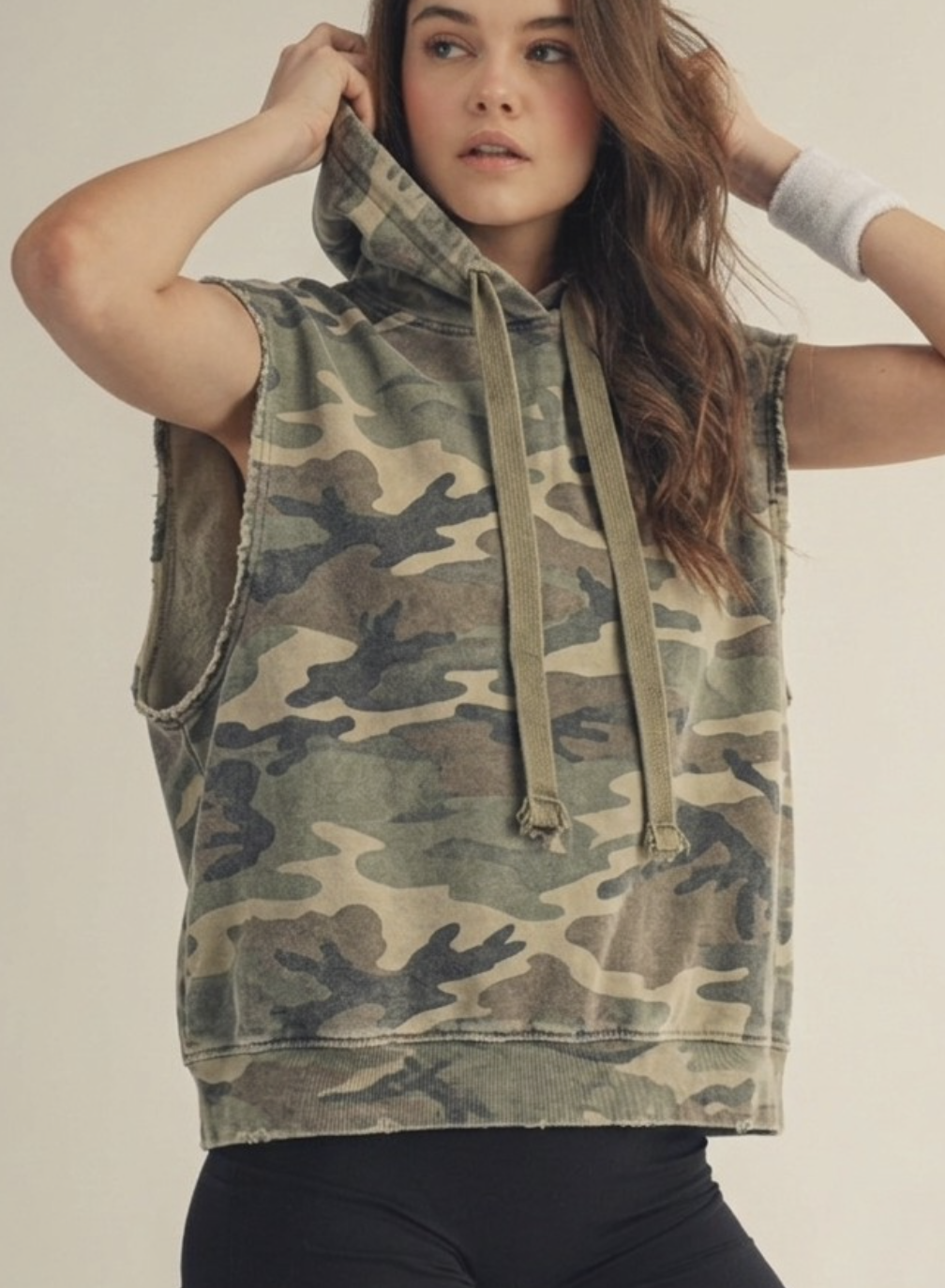 A woman wearing a LovLov sleeveless camouflage hoodie with drawstrings, photographed against a plain light beige studio background.