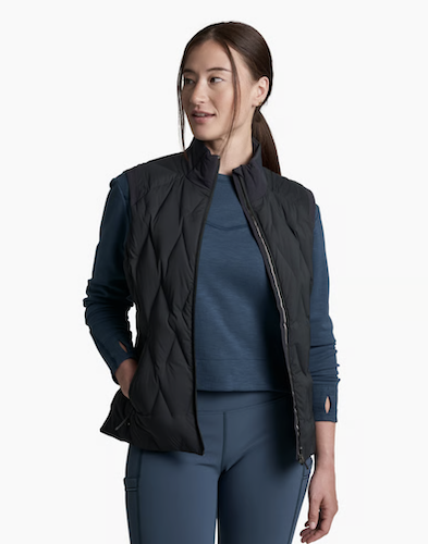 A woman modeling a black Kuhl Firestorm quilted sleeveless puffer vest over a dark blue long-sleeve top and matching blue leggings, set against a plain white studio background.