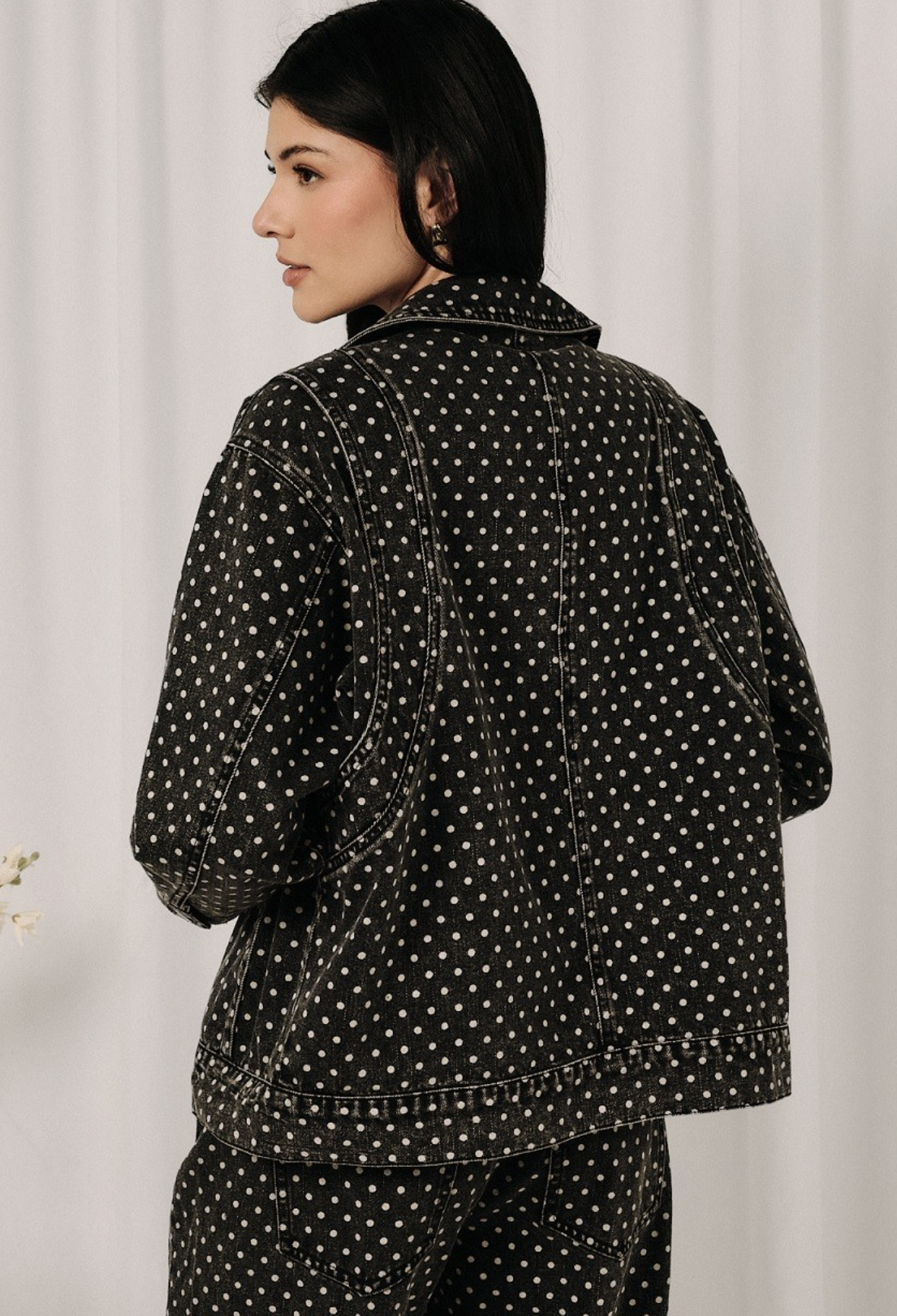 A woman shown from the back wearing a black denim jacket and matching pants set, both featuring a white polka dot pattern, against a light neutral curtain background.