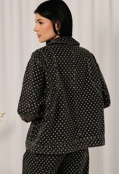 A woman shown from the back wearing a black denim jacket and matching pants set, both featuring a white polka dot pattern, against a light neutral curtain background.