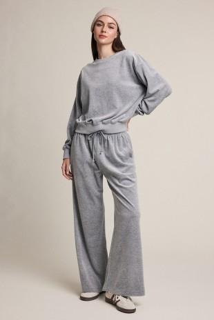 A woman modeling a light gray two-piece lounge set consisting of a long-sleeve sweatshirt and matching wide-leg sweatpants, paired with a beige beanie and white sneakers, against a solid light beige background.