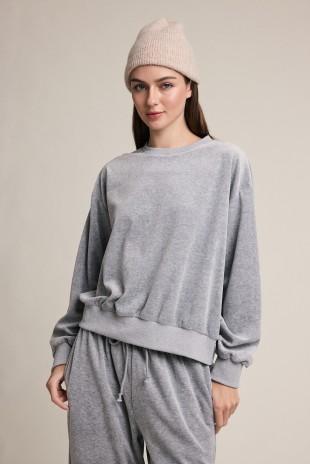 A woman wearing a light gray heathered Raisonnel sweatshirt and matching sweatpants with a beige knit beanie, standing against a plain beige studio background.