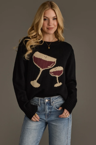 A woman wearing a black knit sweater with a large espresso martini graphic on the front, paired with light blue jeans, standing against a solid dark gray studio background.