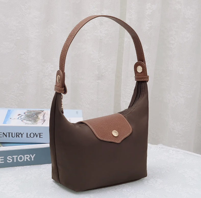 A brown nylon shoulder bag with a brown leather handle and a matching brown flap pocket, styled on a white textured surface next to books against a white curtain backdrop.