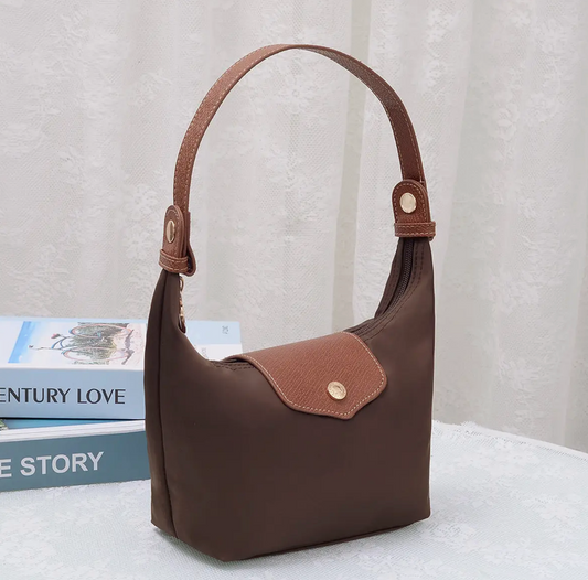 A brown nylon shoulder bag with a brown leather handle and a matching brown flap pocket, styled on a white textured surface next to books against a white curtain backdrop.