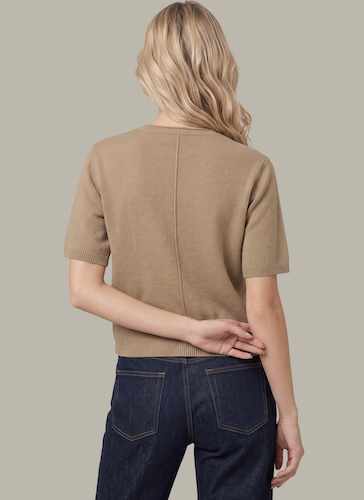 A woman shown from the back wearing a short-sleeve knit top in a warm tan/camel color with dark blue jeans, against a plain light gray studio background.