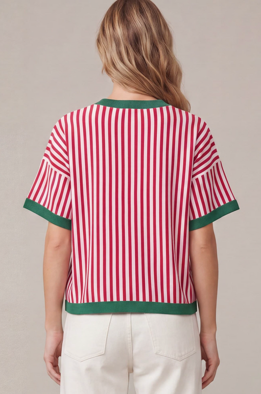 A woman viewed from the back wearing a short-sleeve top with red and white vertical stripes and green trim, paired with off-white pants, against a plain beige studio background.