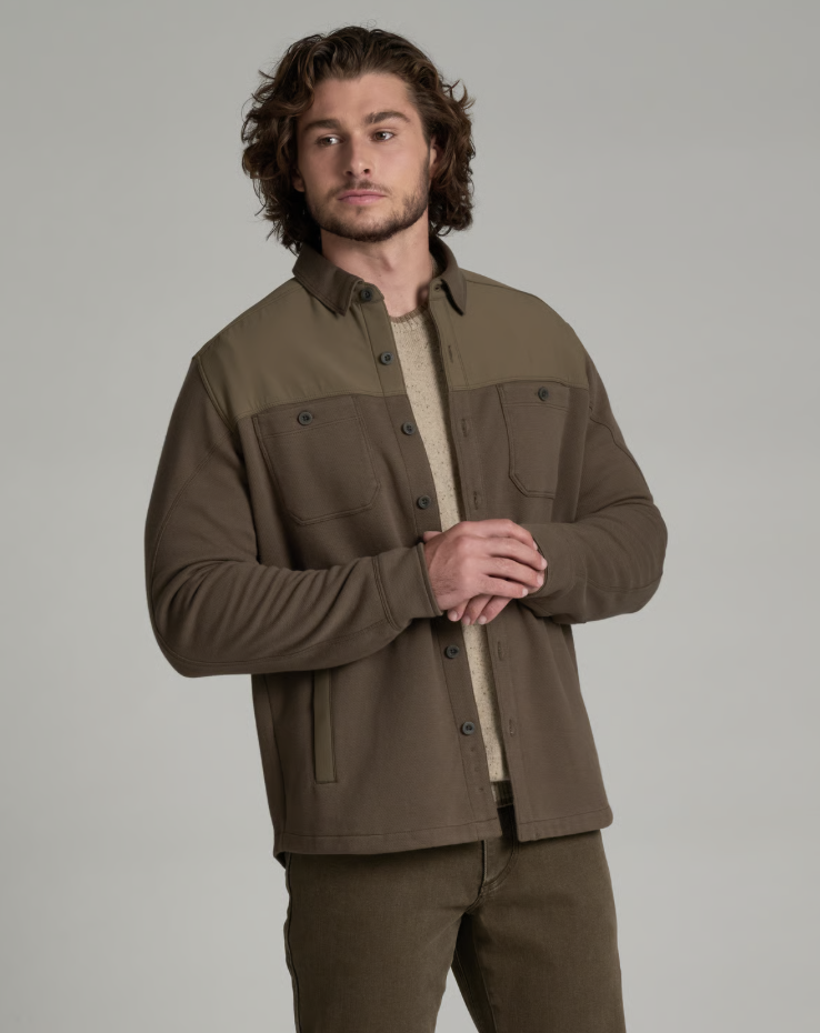 A man wearing a brown and olive green color-blocked Kuhl Relentr button-up overshirt over a beige knit sweater and matching brown pants, photographed against a solid light gray studio background.