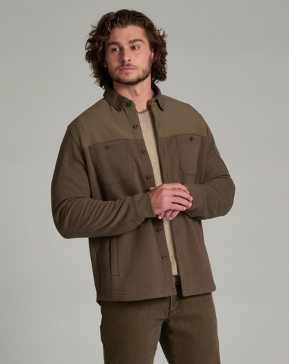 A man wearing a brown and olive green color-blocked Kuhl Relentr button-up overshirt over a beige knit sweater and matching brown pants, photographed against a solid light gray studio background.