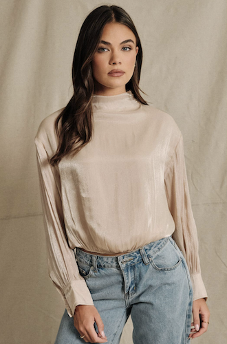 A woman modeling a long-sleeve, high-neck satin blouse in a light beige color, paired with light-wash blue jeans, set against a neutral beige fabric backdrop.
