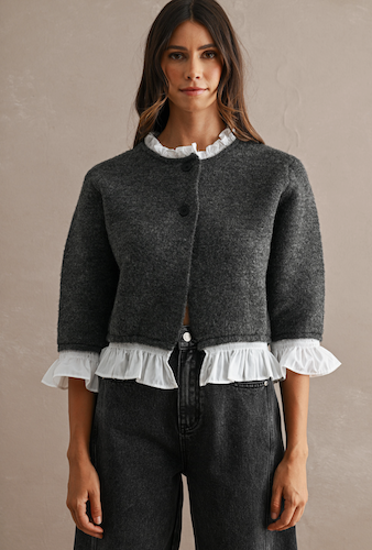 A woman wearing a dark gray cropped cardigan with white ruffled trim at the collar, cuffs, and hem, paired with dark gray jeans, against a neutral textured beige background.