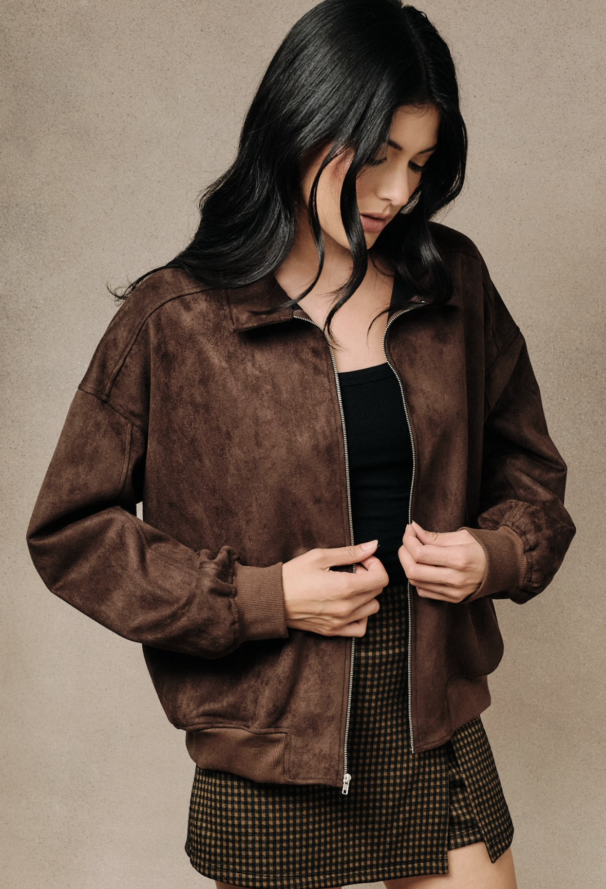 A woman wearing a Brown faux‑suede bomber jacket with modern silhouette and lightweight drape, standing against a neutral beige studio background.