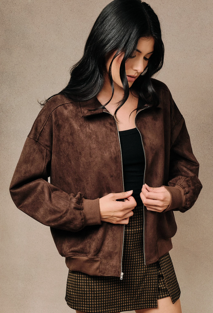 A woman wearing a Brown faux‑suede bomber jacket with modern silhouette and lightweight drape, standing against a neutral beige studio background.