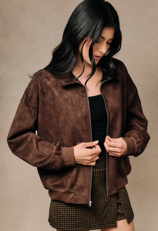 A woman wearing a Brown faux‑suede bomber jacket with modern silhouette and lightweight drape, standing against a neutral beige studio background.