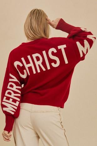A woman shown from the back wearing a red knit sweater with large white 'MERRY CHRISTMAS' lettering across the back and sleeves, paired with light beige pants, against a plain beige background.