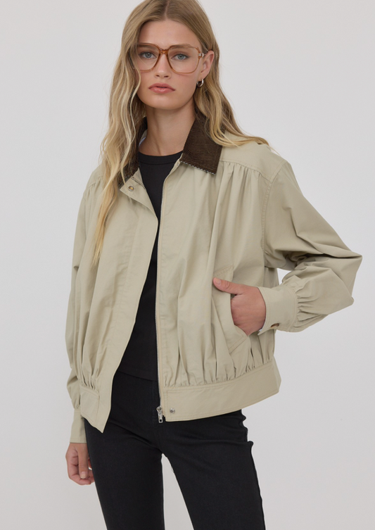 A woman wearing a light beige utility jacket with a dark brown collar, styled over a black top and black pants, photographed against a plain light gray studio background.